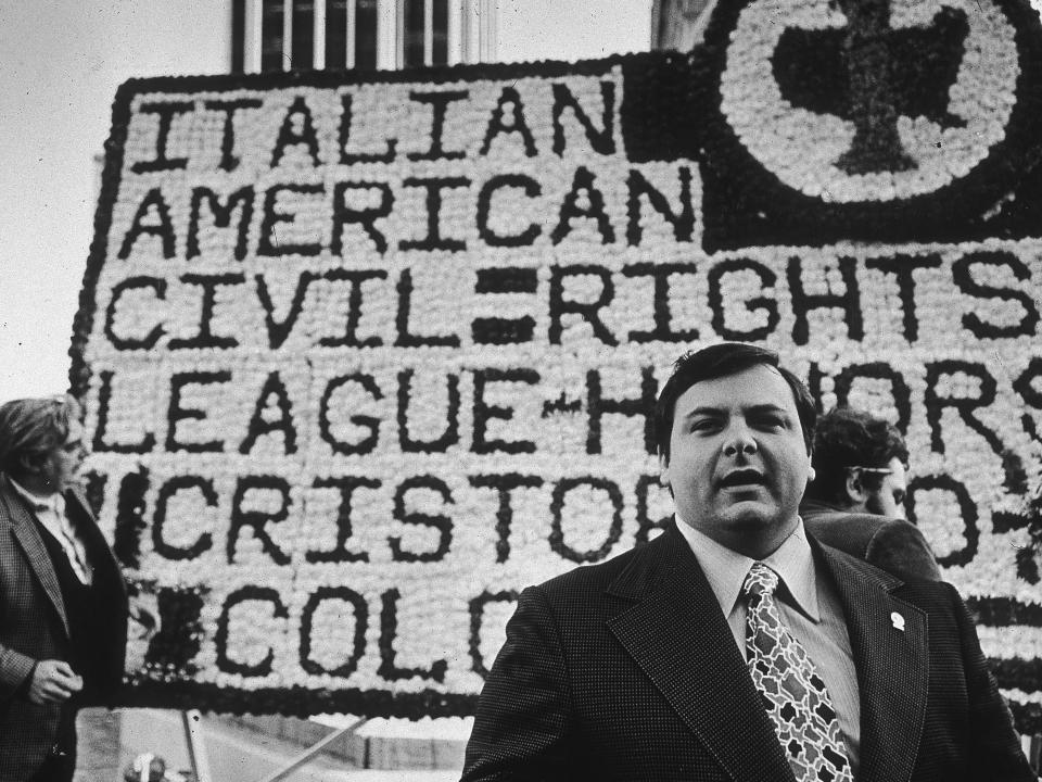 Colombo in a suit in front of a large floral tribute for the Italian-American Civil RIghts League.