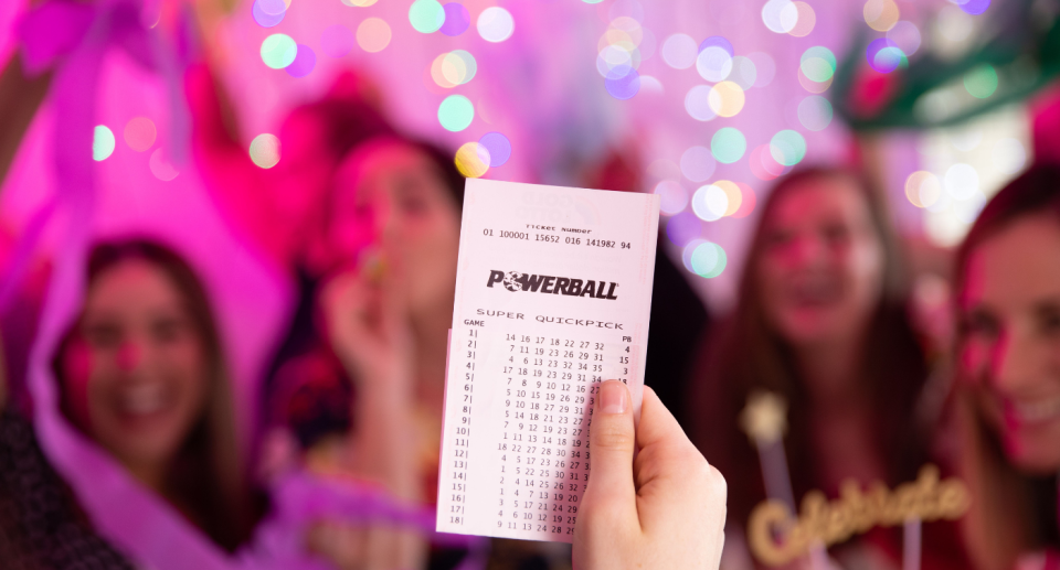 A person holding up a Powerball ticket with people celebrating in the background.