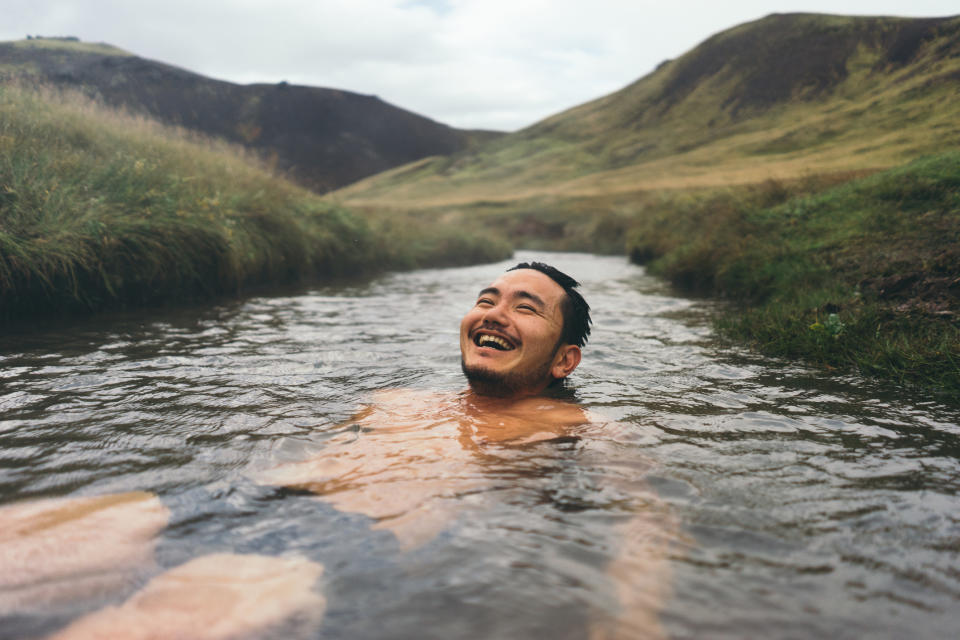 A person smiles joyfully while floating in a stream surrounded by grassy hills. The image conveys a sense of relaxation and happiness in nature