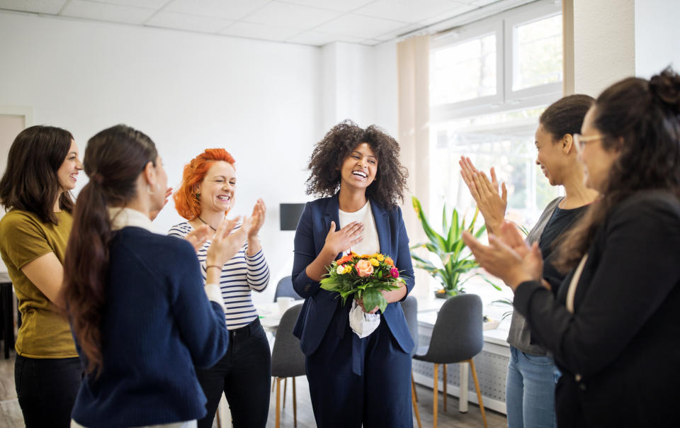 Pictured: Smiling woman with flowers laughs as colleagues applaud her promotion. Image: Getty
