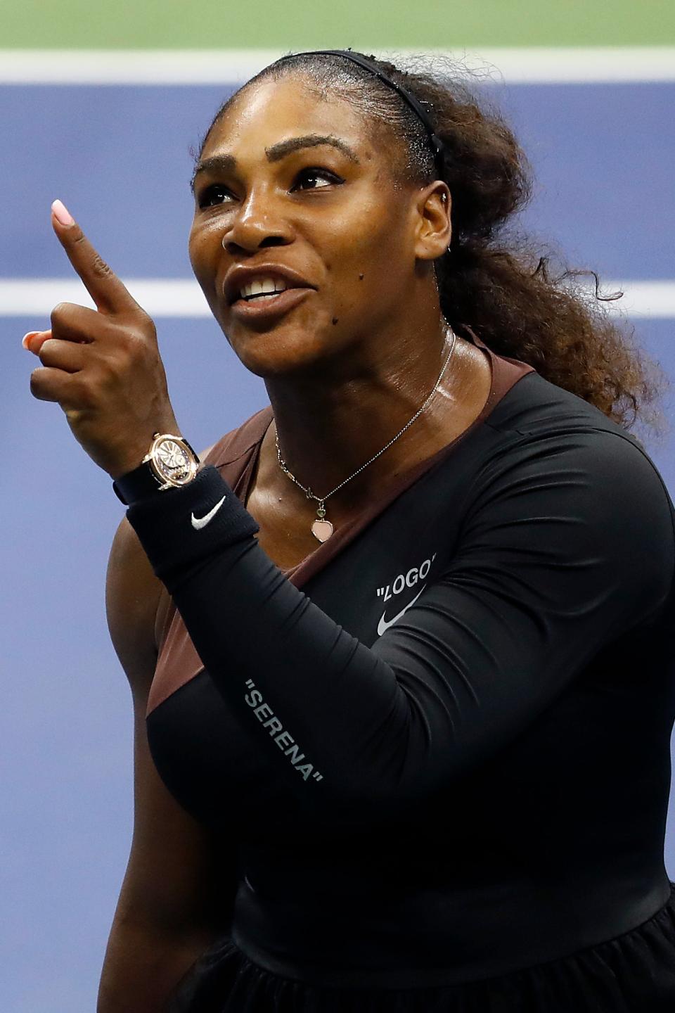 Serena Williams in action on a tennis court, gesturing upwards with her index finger