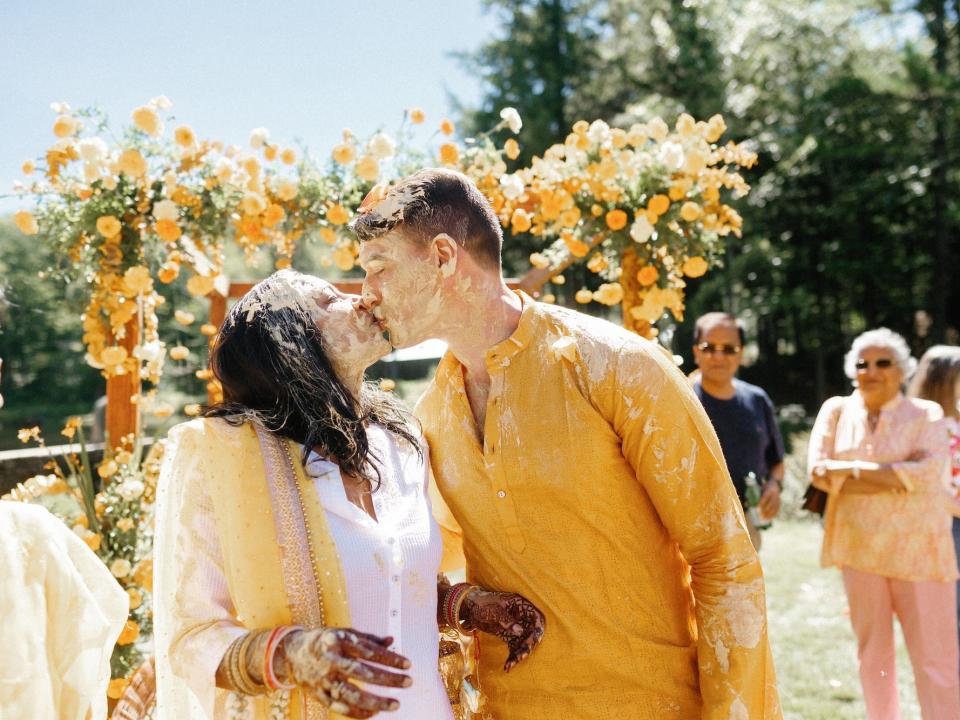 A bride and groom kiss in front of floral archway during their haldi ceremony.