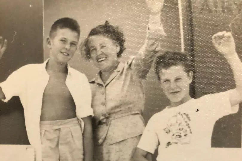 Black and white photo of John Teasdale (L) and Christopher Lawrence (R) aged 11 with John's mother at London Airport in 1959 posing with one hand raised holding bags that say "Airways" on, in faux aeroplane door