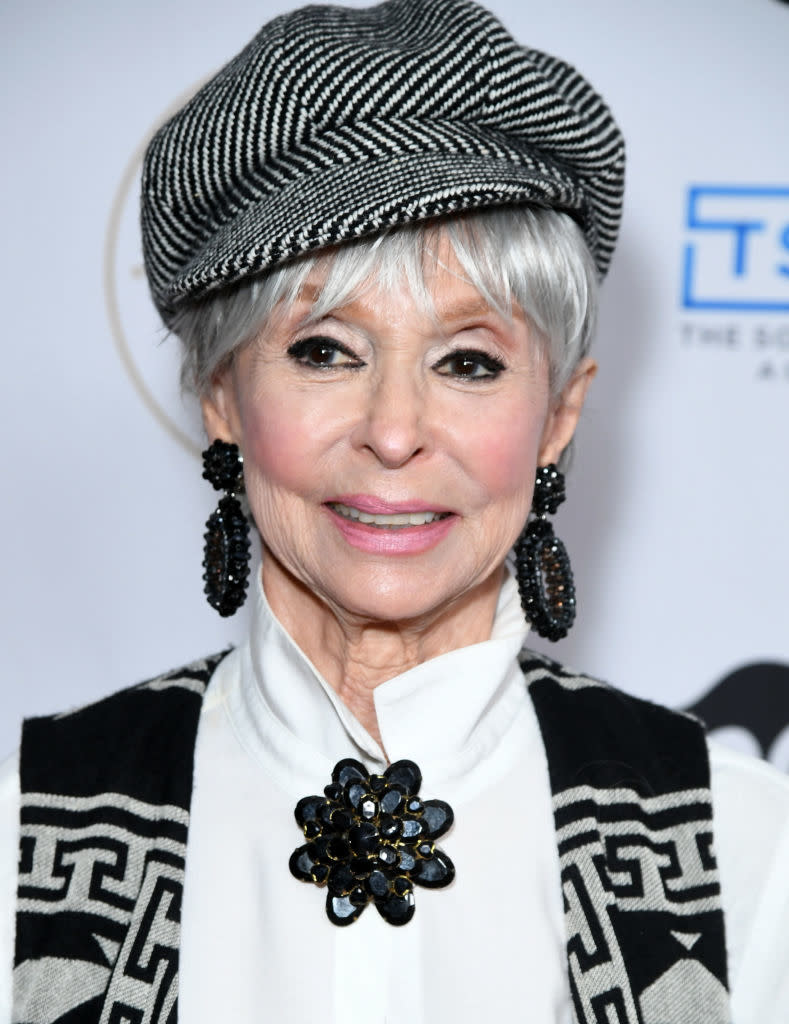 Close-up of Rita wearing a cap, blouse and vest, and large broach and earrings at a media event