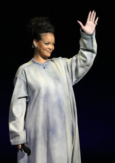 Rihanna in a long jean dress holding up her left hand while standing on a stage