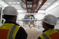 Workers, foreground, stand Wednesday, Oct. 11, 2023, in an area that features a life-sized representation, behind, of a device that will use magnets to create the conditions for fusion to happen, at Commonwealth Fusion Systems, in Devens, Mass. Commonwealth is trying to create fusion inside what's called a tokamak. Nuclear fusion, which would be a new source of carbon-free energy, melds two hydrogen atoms together to produce a helium atom and a lot of energy. (AP Photo/Steven Senne)