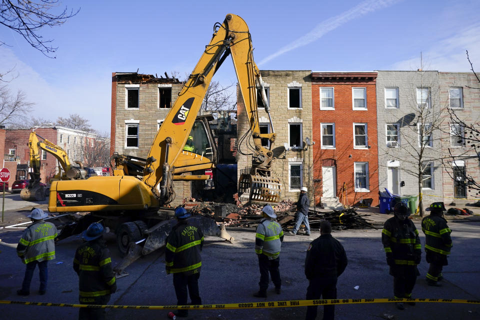 Fire officials and a crew look on as an excavator is used to pull debris off a building during efforts to retrieve the body of a deceased firefighter caught in the building's collapse while battling a two-alarm fire at the vacant row home, Monday, Jan. 24, 2022, in Baltimore. Officials said several firefighters died during the blaze. (AP Photo/Julio Cortez)