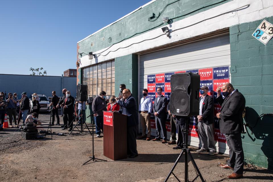 Rudy Giuliani speaks at a lectern in an unpaved parking lot at a landscaping business in Pennsylvania with some people behind him