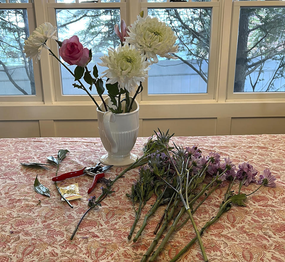 This May 1, 2024 image provided by Jessica Damiano shows a supermarket bouquet of three white chrysanthemums, a lily stem and a single rose in a decorative pitcher, along with garden pruners and a packet of flower-preserving granules on Long Island, New York. Proper preparation and the addition of greenery and "filler" flowers can transform a simple bouquet into a lovely arrangement. (Jessica Damiano via AP)