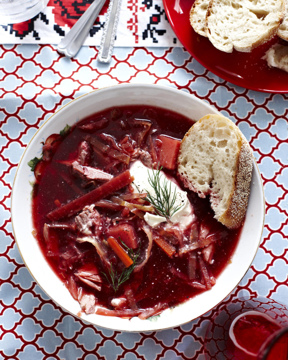 A bowl of borscht soup with a slice of bread and a dollop of sour cream, garnished with dill, on a patterned tablecloth