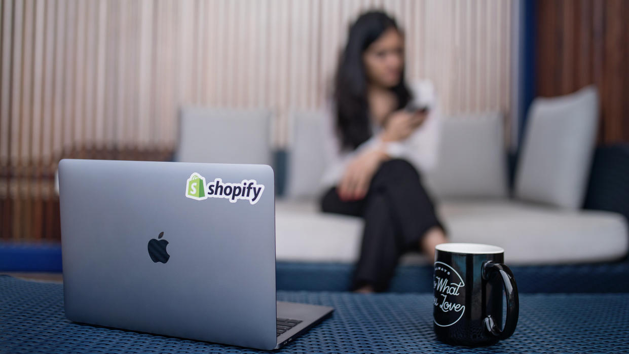  Photograph of laptop with Shopify sticker, on table beside coffee mug, with woman on her phone out of focus in background. 