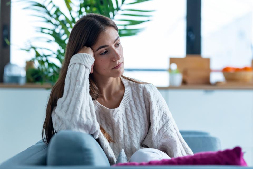 A woman with long brown hair sits on a couch, resting her head on her hand, looking pensive. She wears a cozy, knit sweater and is surrounded by home decor items