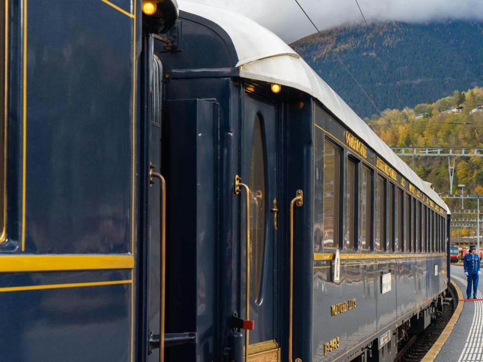 A navy blue train with gold trimmings stopped at a platform with mountains in the background