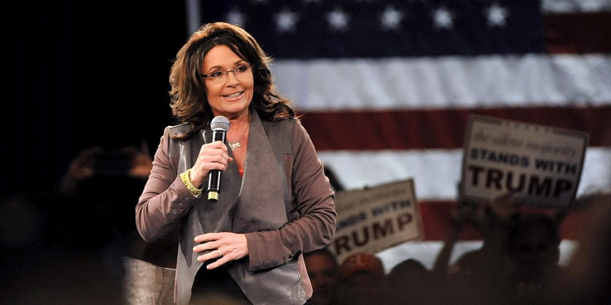 Sarah Pain holds a microphone on stage in front of an American flag and Trump signs