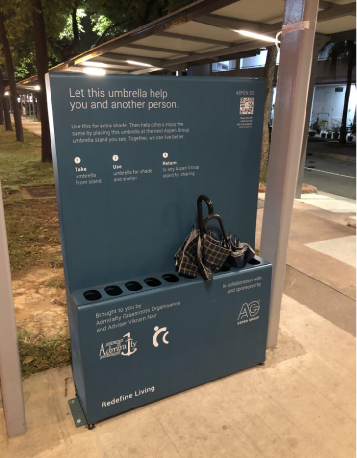 A bin on a street corner hold umbrellas, with a sign that says "let this umbrella help you and another person"