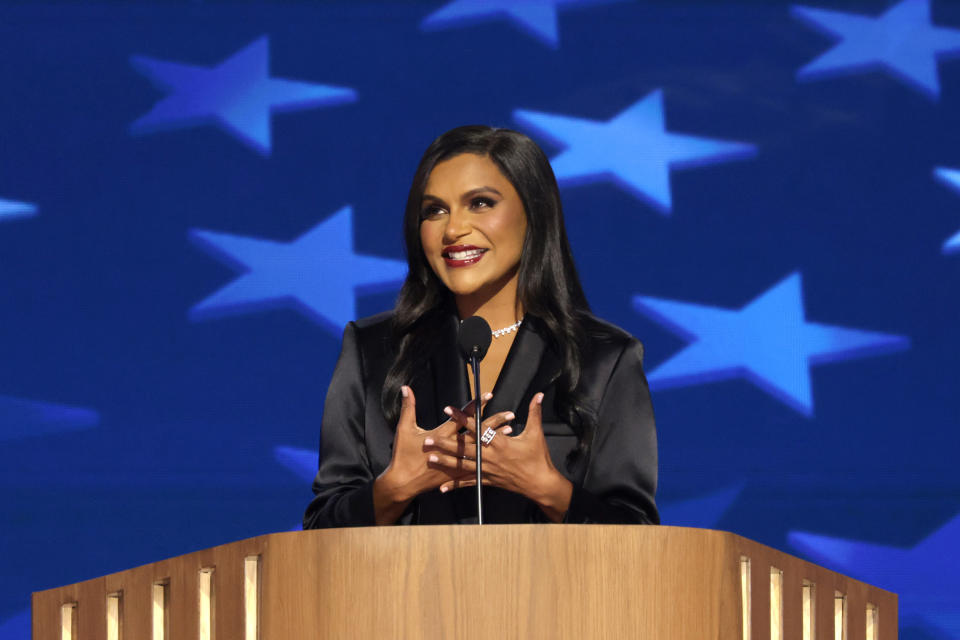 Mindy Kaling spricht auf einem Podium und gestikuliert mit ihren Händen vor einem Hintergrund aus Sternenmotiven. Sie trägt ein schlichtes, elegantes Outfit