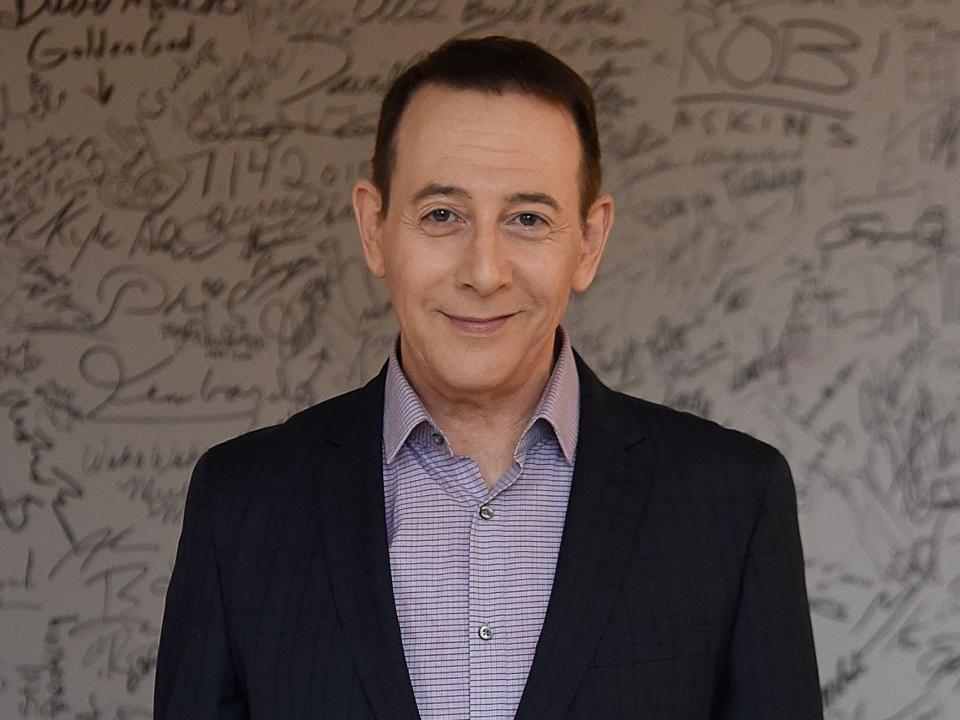 Paul Reubens in a dark-blue suit with a light blue shirt in front of white wall covered in handwritten messages