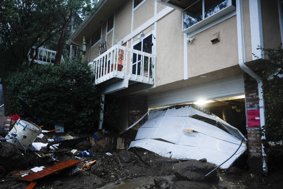 A garage door is damaged next to debris strewn about a house.