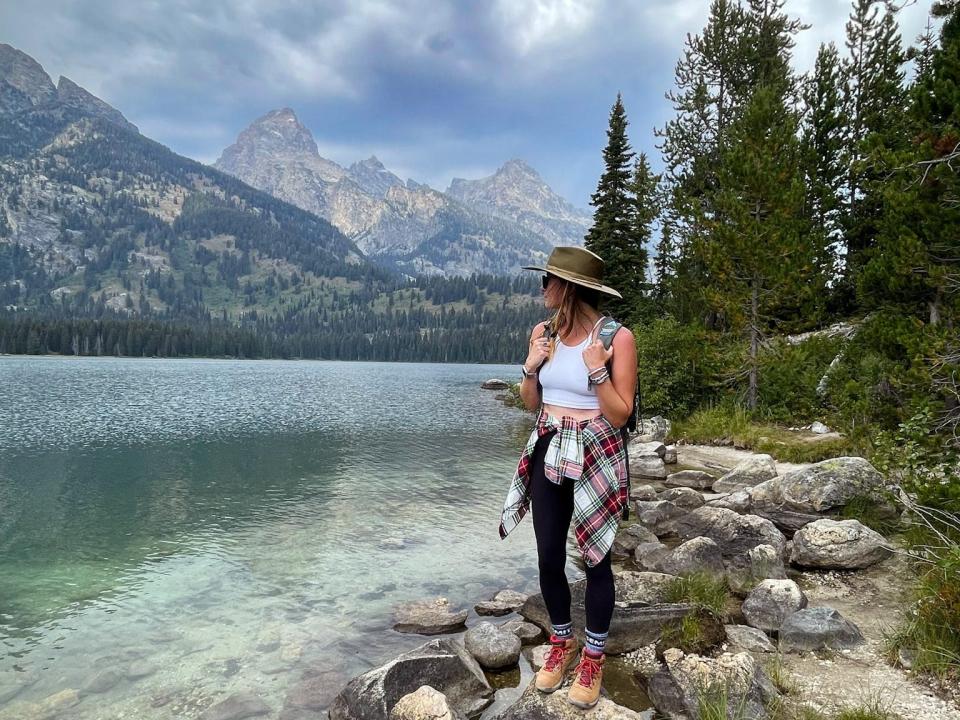 Emily, wearing a white shirt, black leggings, a hat, a backpack, and a flannel around her waist, stands on a rock looking out at clear water, mountains, and trees.