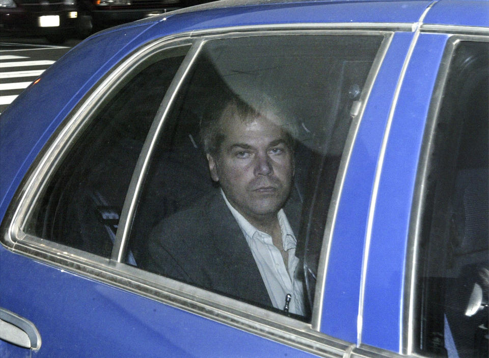 John Hinckley Jr., wearing a suit jacket and unbuttoned-at-the-top shirt, looks out the window of the backseat of a blue car.