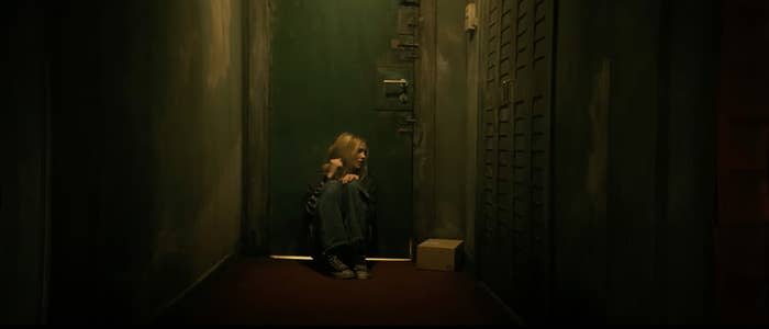 A young woman with blonde hair sits on a hallway floor near a door, looking scared and holding her knees close to her chest