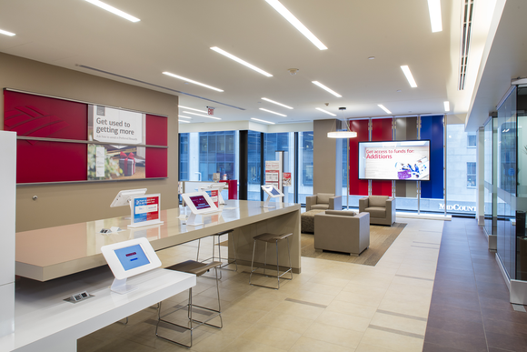 Lobby of a modern Bank of America branch, with white counters and tablets on top and comfy beige chairs in the background.