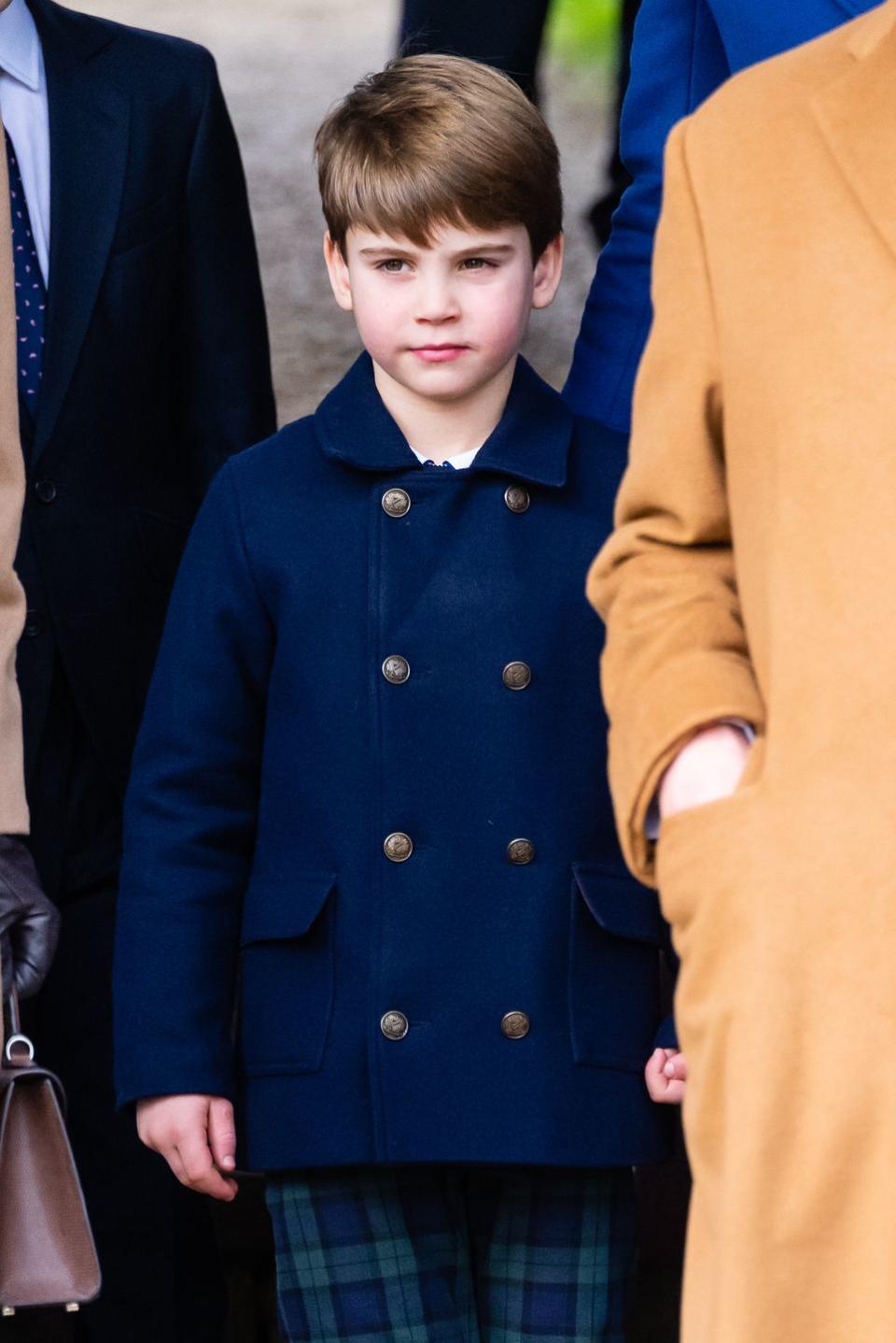 prince louis of wales stands in a blue peacoat and blue and green plaid pants among other people