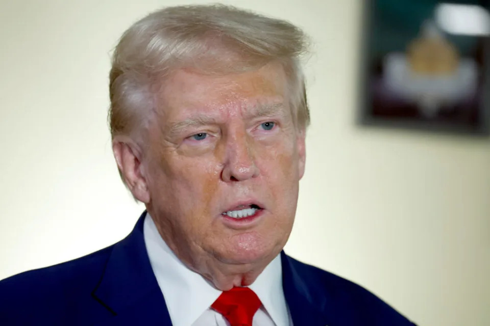 Donald Trump speaking, wearing a suit and red tie. Background is blurred