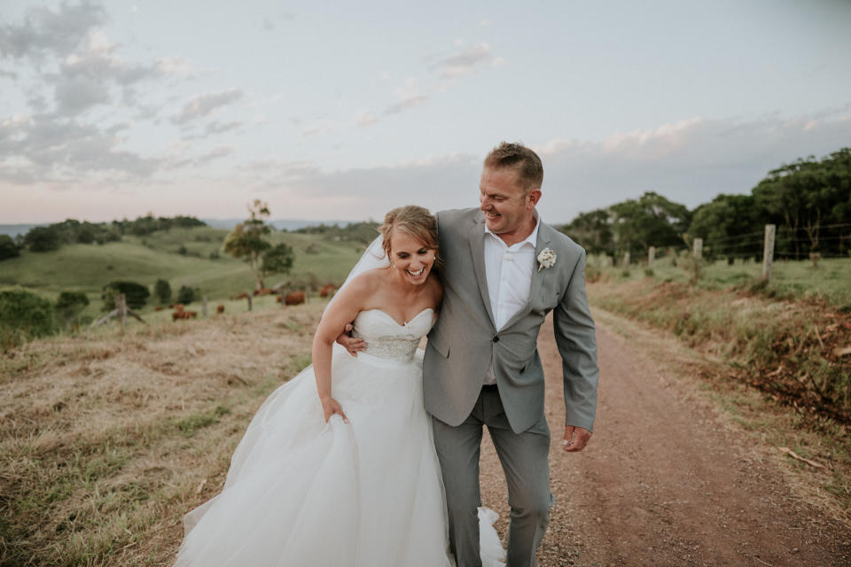 The bride and groom stand together laughing on a dirt road with green hills and a pale blue, partly cloudy sky behind them. The groom has his arm wrapped around the bride's waist as she holds her skirt in one hand. (Photo: <a href="https://www.jamesday.com.au/" target="_blank">James Day Photography</a>)