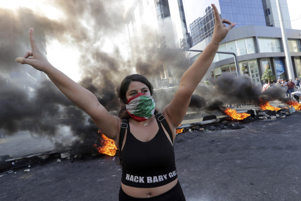 An anti-government protester, her face wrapped with a Lebanese flag, makes victory signs standing in front of burning tires that block a road in the town of Jal el-Dib, north of Beirut, Lebanon, Wednesday, Nov. 13, 2019. Lebanese protesters blocked major highways with burning tires and roadblocks on Wednesday, saying they will remain in the streets despite the president's appeal for them to go home. (AP Photo/Hassan Ammar)