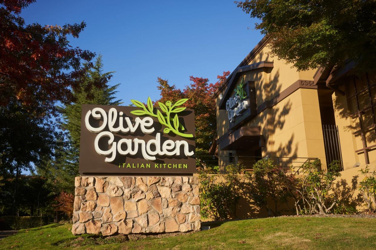 Sign in the foreground with garden area and with exterior in background of an Olive Garden restaurant, Lake Oswego, Oregon with a blue sky in the background during late summer