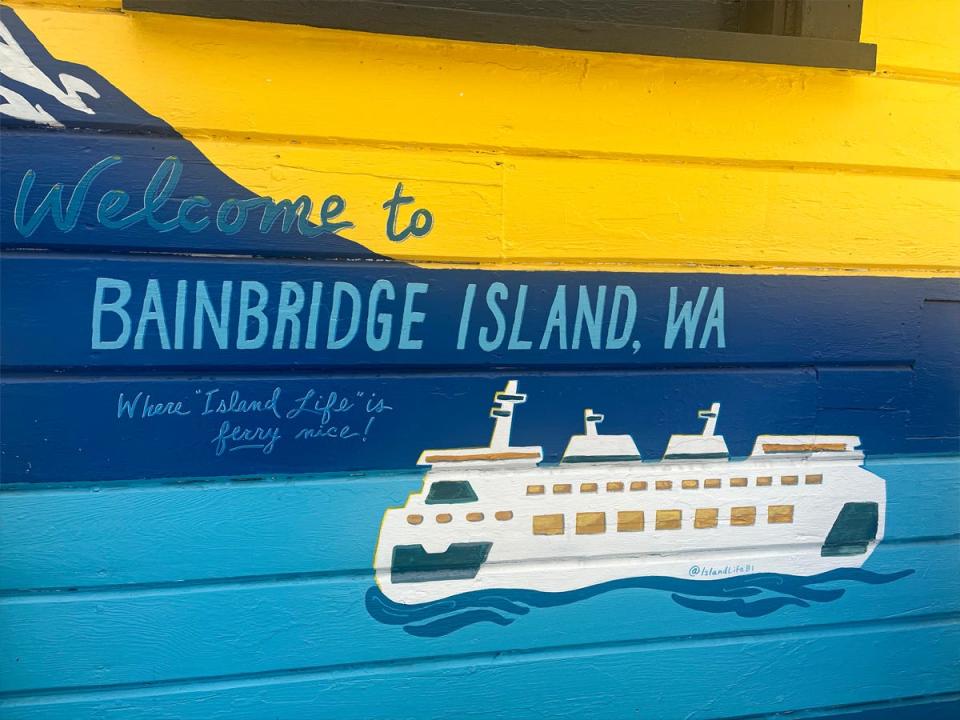 A painted ferry with a yellow, dark-blue, and lighter blue on the side of a building with painted letters spelling out "Welcome to Bainbridge Island, WA"