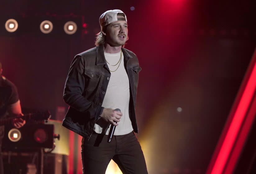 Morgan Wallen performing on stage, holding mic, wearing white hat backwards, black jacket, white t shirt and dark pants.