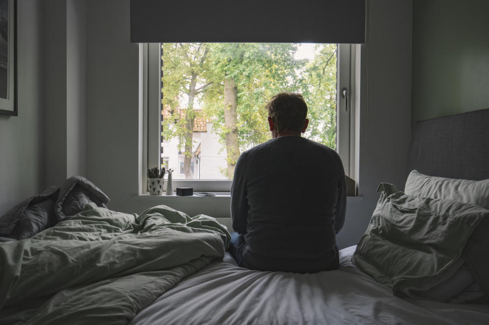 Person sits on the edge of an unmade bed, looking out of a window at trees. Bedroom setting suggests contemplation or reflection