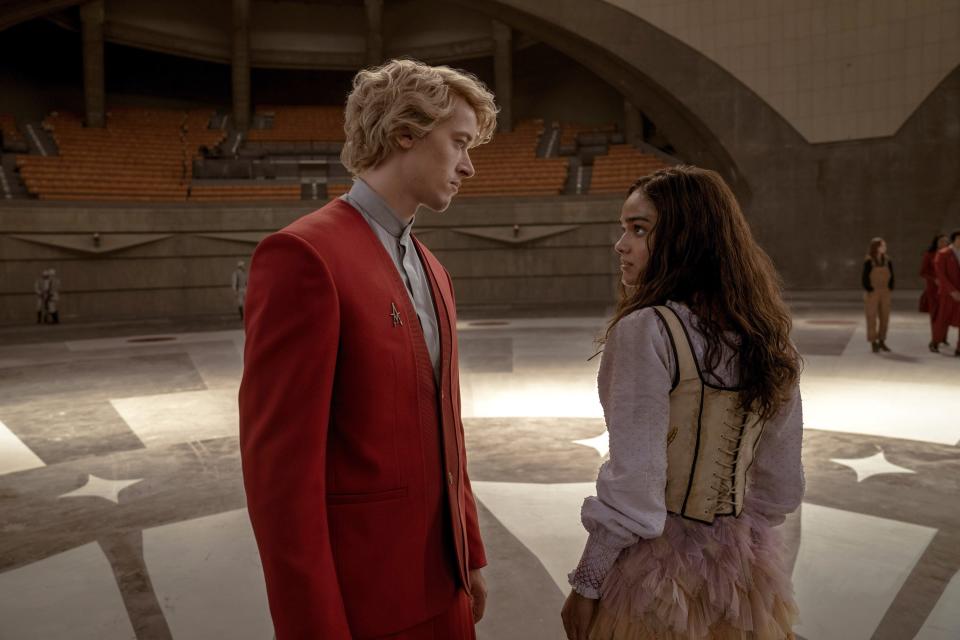 Tom Blyth in a red suit and Rachel Zegler in a period costume stand facing each other in a large, indoor arena