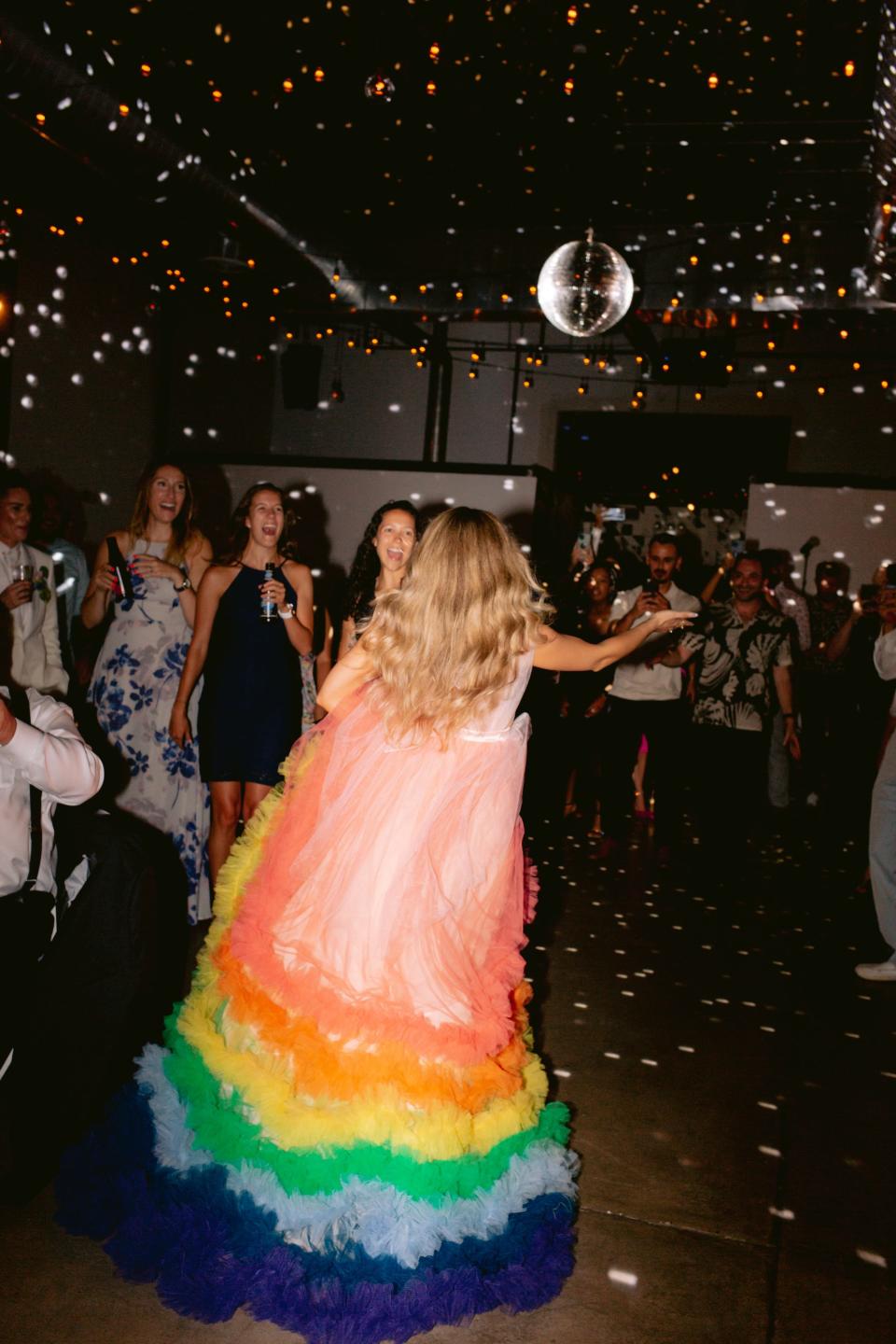 A bride in a rainbow, ruffled dress walks towards her wedding guests.