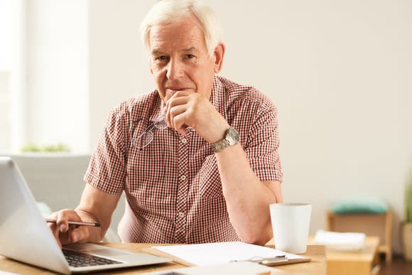 A gray-haired man sits at a desk in front of a laptop, holding his glasses in one hand.