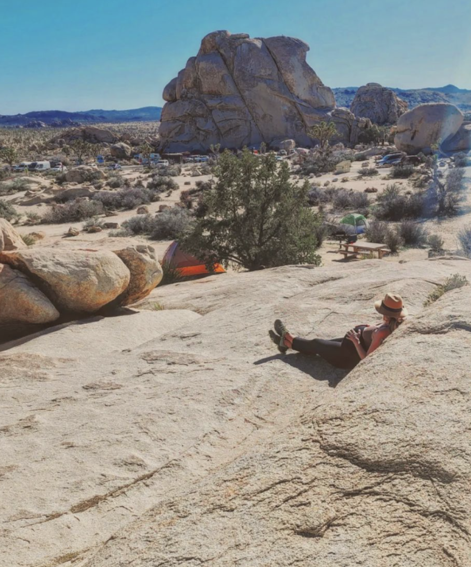 Person in a hat resting on a rocky slope with boulders and desert vegetation in the background