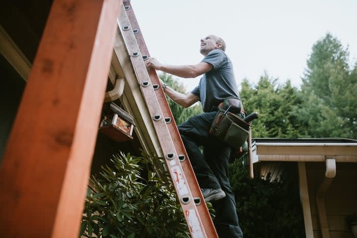 Man with tool belt climbing ladder to roof.