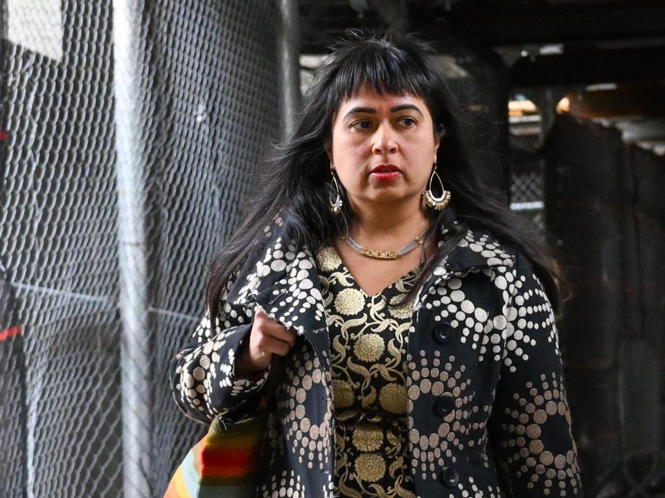 Priya Chaudhry walking past a chain-link fence while wearing a black and white jacket, large golden earrings, and a multicolored handbag.