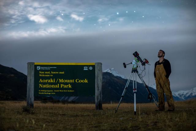 Brian Bezalel stand in front of sign for Aoraki / Mount Cook National Park standing next to a telescope.