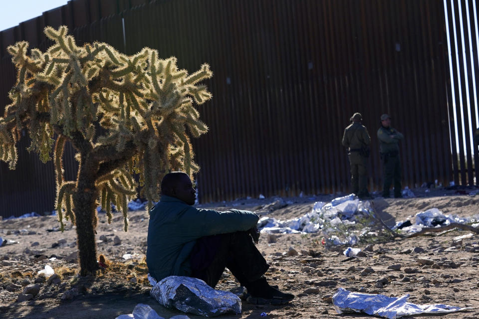 A migrant sits in the shade provided by a desert cholla cactus as he joins hundreds of migrants gathering along the border Tuesday, Dec. 5, 2023, in Lukeville, Ariz. The U.S. Border Patrol says it is overwhelmed by a shift in human smuggling routes, with hundreds of migrants from faraway countries like Senegal, Bangladesh and China being dropped in the remote desert area in Arizona. (AP Photo/Ross D. Franklin)