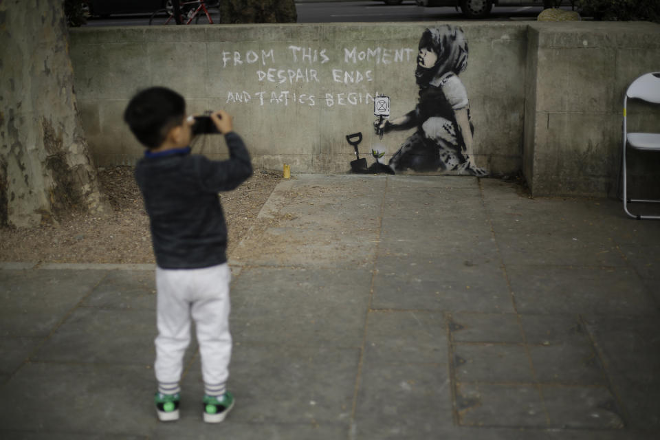 A young boy points a camera he was playing with towards a new piece of street art believed to be by street artist Banksy on a wall where Extinction Rebellion climate protesters had set up a camp in Marble Arch, London, Friday, April 26, 2019. Extinction Rebellion ended its remaining blockades in London on Thursday evening with a closing ceremony, after disrupting the British capital for 10 days. The non-violent protest group is seeking negotiations with the government on its demand to make slowing climate change a top priority. (AP Photo/Matt Dunham)