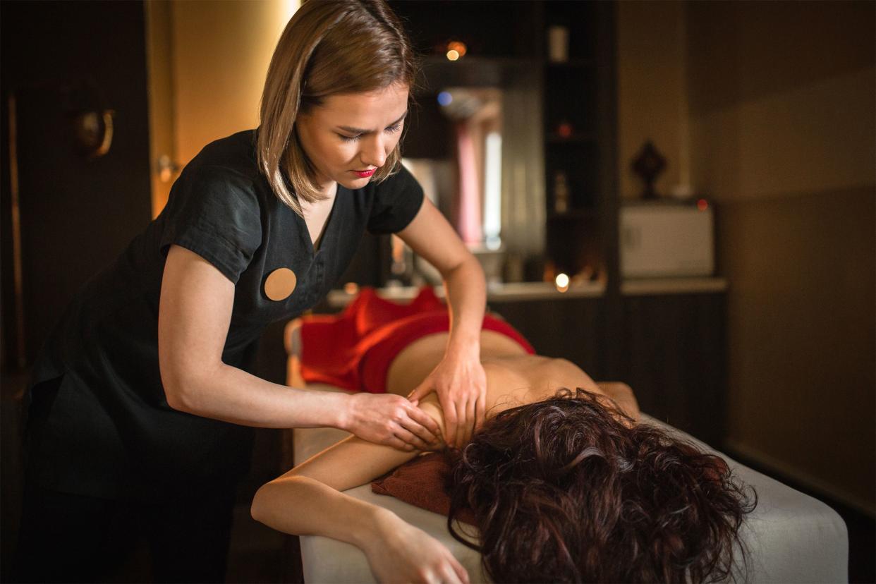 Female massage therapist giving a massage to a woman lying face down on a massage table, therapist and head of woman is in the foreground, candle-lit massage room blurred in the background