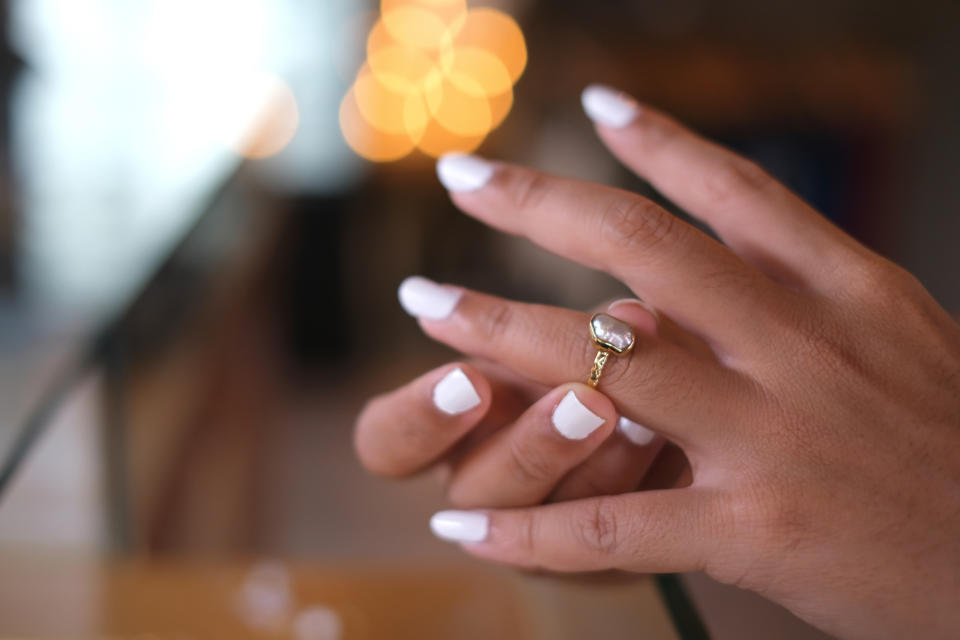 A close-up of a woman's hand with white nail polish, wearing a diamond ring on her ring finger