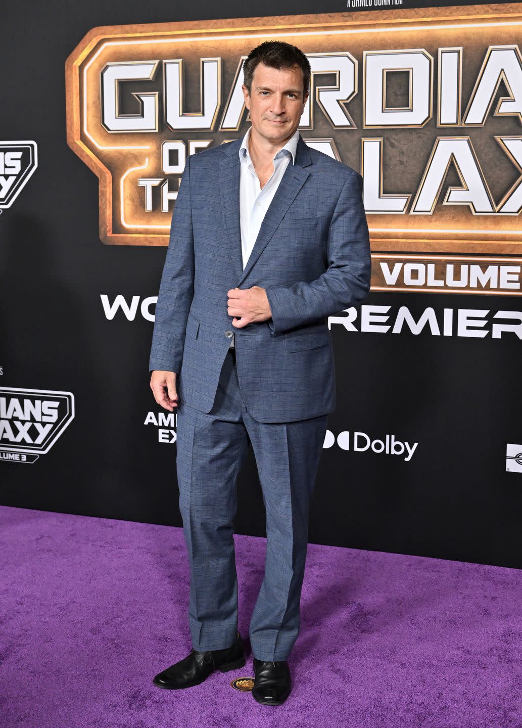 nathan fillion wearing a white collared shirt and a navy blue suit, standing in front of a background with the words guardians of the galaxy volume 3 on it at the world premiere of guardians of the galaxy volume 3 in april 2023