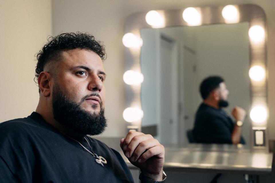 Mo Amer looks to his right as he sits near a mirror with lights around it. His reflection is seen.