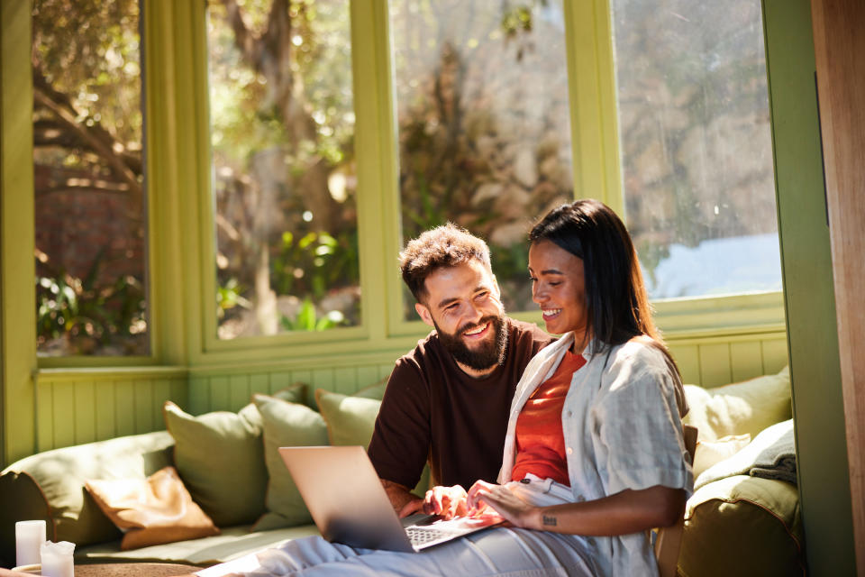 Laughing young couple sitting together in their sunny living room at home and working on a laptop