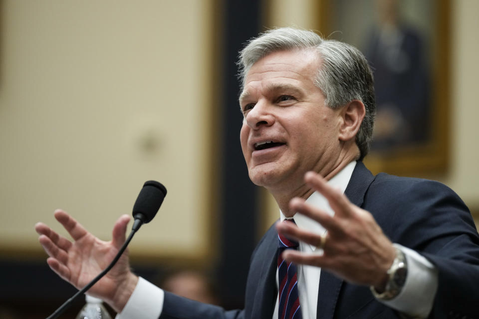 FBI Director Christopher Wray, at the microphone, gestures expansively, with a look of great good humor. 