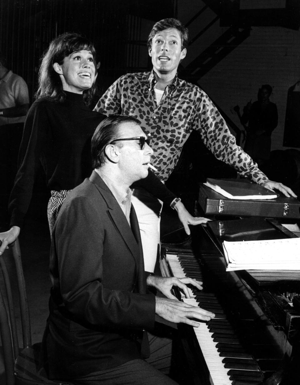 Man in sunglasses playing piano, a woman and another man singing behind him in a casual setting